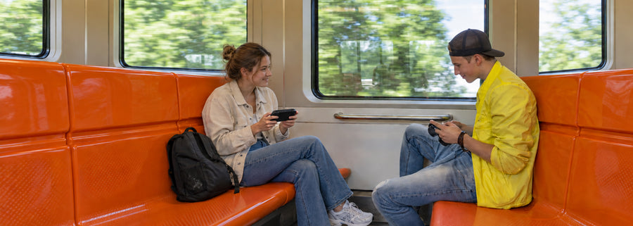 Two passengers playing AISLPC Pocket AIS handheld game consoles while sitting on a train, highlighting portable gaming for travel.